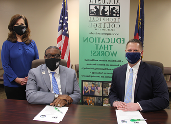 Caucasian male wearing a blue face mask, blue suit jacket, white collared shirt, light blue tie, hands folded while sitting down at a table, white paper in front of him with a green pen; African American male wearing a black Augusta Tech branded face mask, light gray suit, silver Augusta Tech lapel pin, white collared shirt, blue/purple tie, silver watch, hands folded white sitting at a brown table, white papers on brown table in front of him with a green pen; Caucasian female standing wearing a black Augusta Tech branded face mask, blue shirt, hands folded together, black pants, background features an American flag, the state of Georgia flag, large retractable banner showing old Augusta Tech logo, verbiage reads: gqkqu.goudounet.com, Education That Works; bulleted list reads Allied Health Sciences and Nursing, Business and Personal Services, and Engineering Technology, Industrial Technology, and Learning Support, photos underneath the bulleted list.