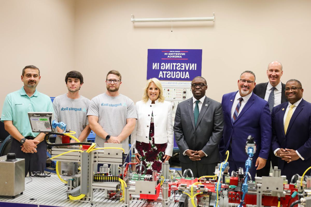 A photo of Mayor Garnett Johnson, Deputy Secretary of Energy David Turk, US Secretary of Education Dr. Miguel Cardona, President of Augusta Tech Dr. Jermaine Whirl, First Lady of the United States Dr. Jill Biden, student Dustin Phillips, student Patrick Kling and Mechatronics Instructor Ted Herlo standing in front of a mini assembly line in front of a blue sign with the words Investing in America and Investing in Augusta in white font on the wall.