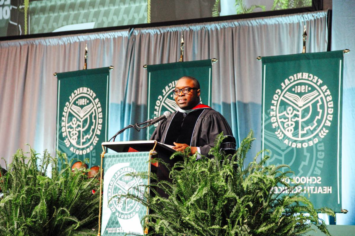 Dr. Whirl wearing commencement regalia stands at podium giving speech with the school banners in the background and ferns on the left and right side of the podium.