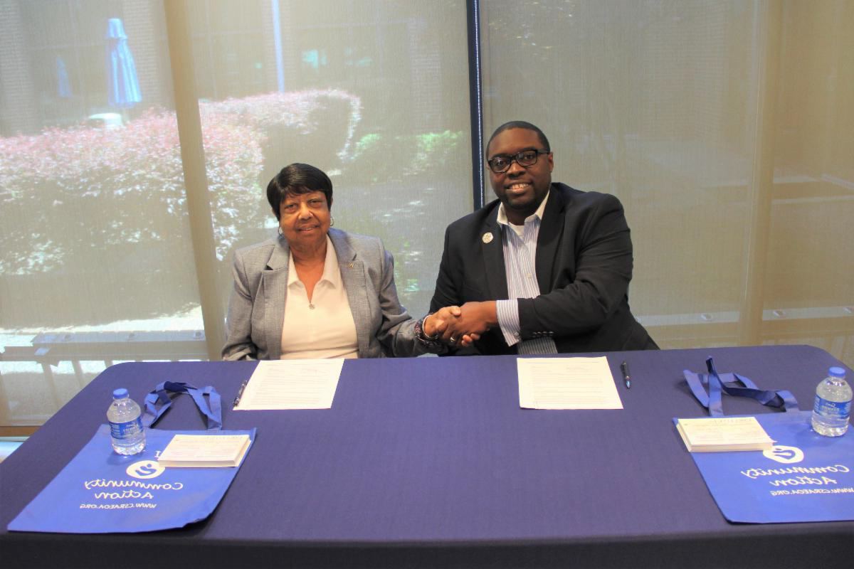 African American male wearing a blue blazer, white collared shirt, silver lapel pin, sitting in a chair shaking hands next to an African American female wearing a gray blazer, white shirt, both are sitting in front of a table with a blue table cloth, on the table are white papers and blue and white bags. The photo is in front of a window, and outside the window are flowers.