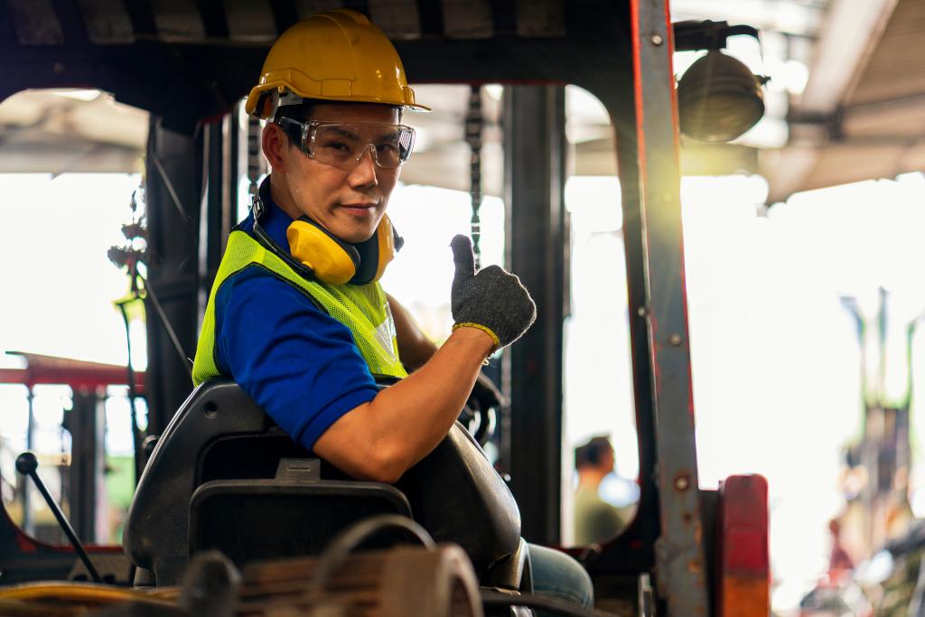 A forklift operator sits in the seat of a forklift wearing a yellow hard hat, safety glasses and yellow safety vest with yellow noise cancelling headphones around his neck.