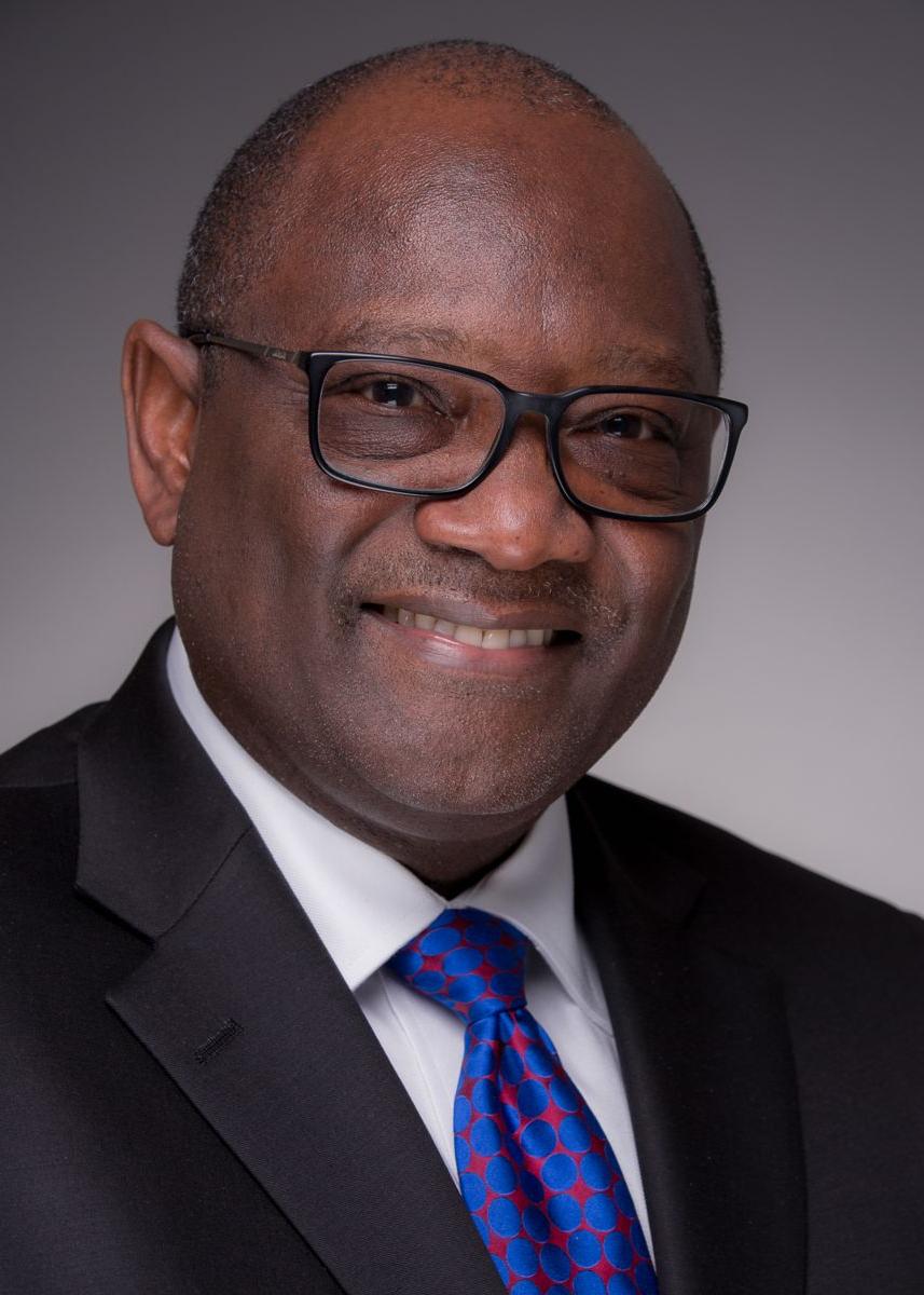Mr. John Richardson, an African American male, smiling wearing black frame glasses, a black suite with a blue tie with a red pattern.