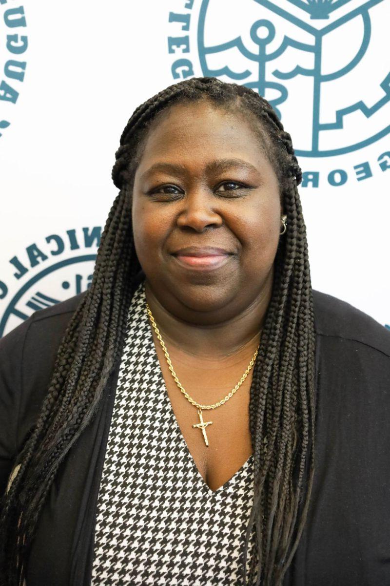 Tiffany Rowe-Thomas, an African American female with long braided hair worn in a half up, half down style smiles at the camera wearing a black and white checkered top and a black sweater against a backdrop of the Augusta Technical College Seal in heritage green on a white background.